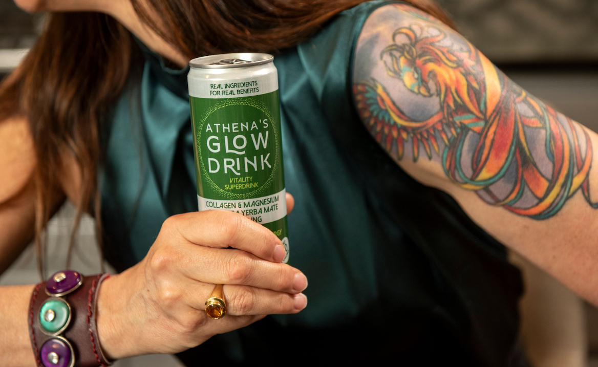 Woman holding a can of Athena's Glow Drink with visible tattoos on arm.