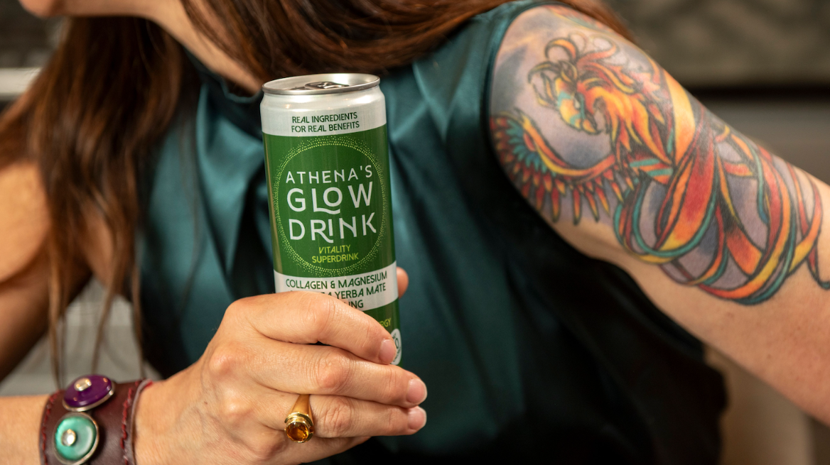 Woman holding a can of Athena's Glow Drink with visible tattoos on arm.