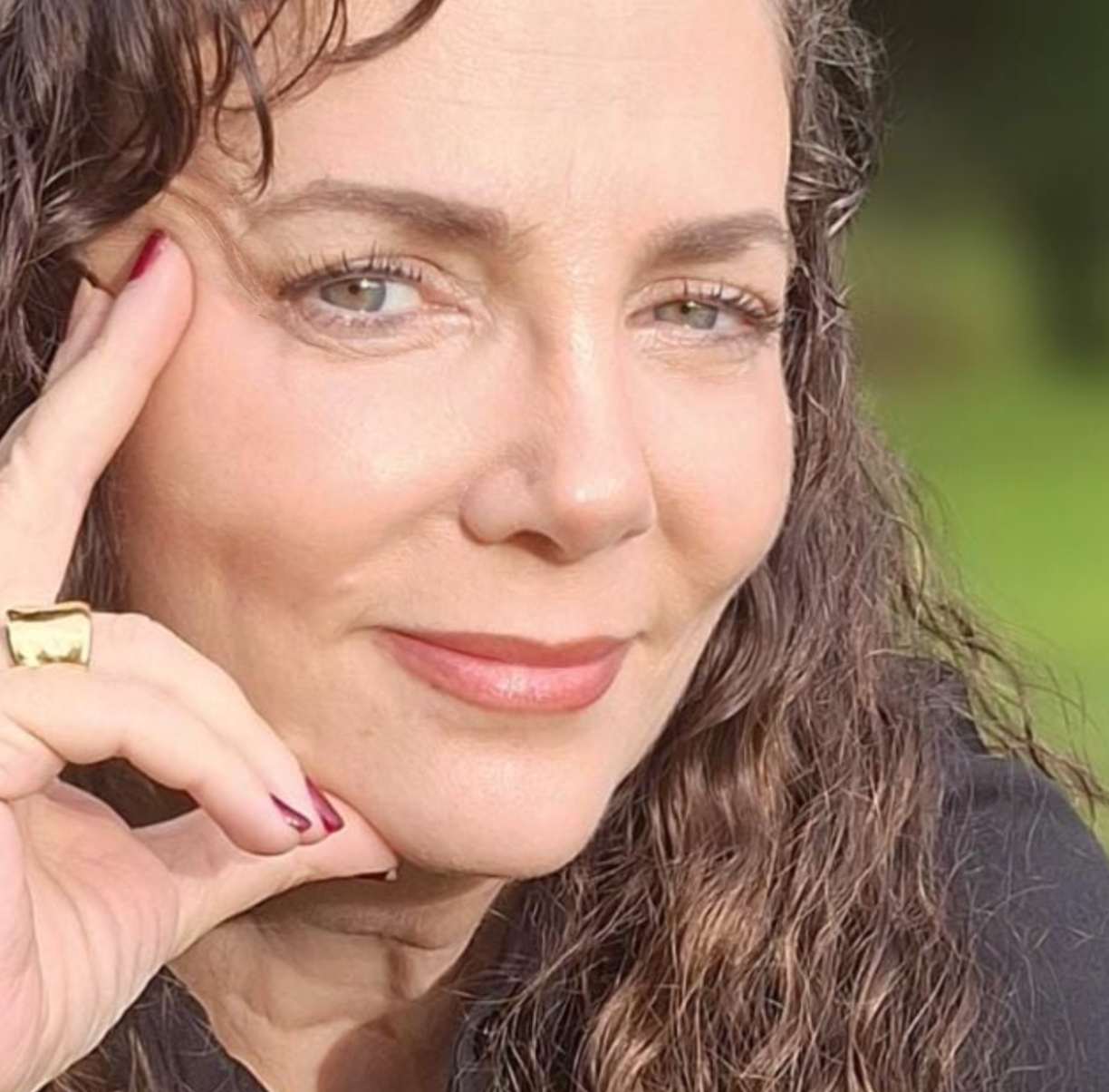 Woman with wavy brown hair and a gold ring on her finger, outdoors.
