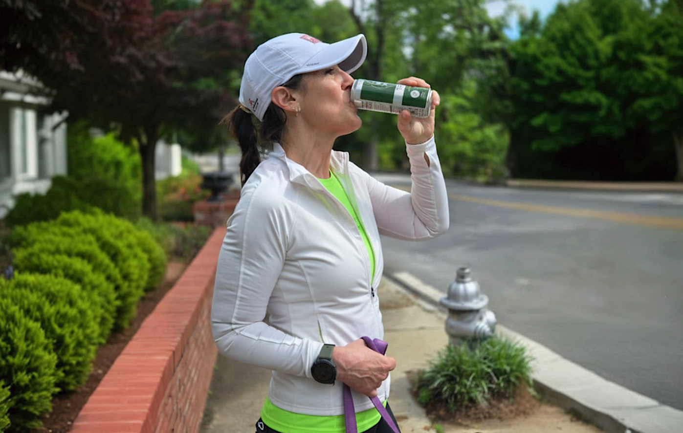 Woman taking a break from running to drink from an Athena's Glow Drink can on a sidewalk.