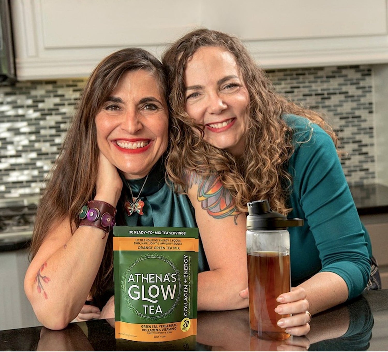 Athena's Glow founders in a kitchen with Athena's Glow Tea packaging and a glass of tea.