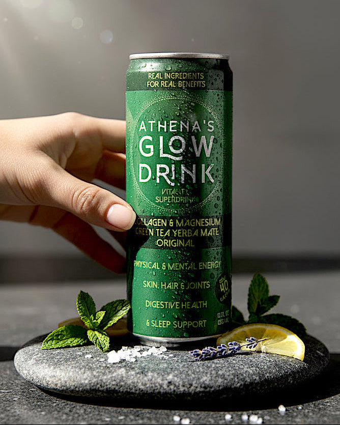 Hand holding a can of 'Athena's Glow Drink' Original with herbs and lemon on a dark surface