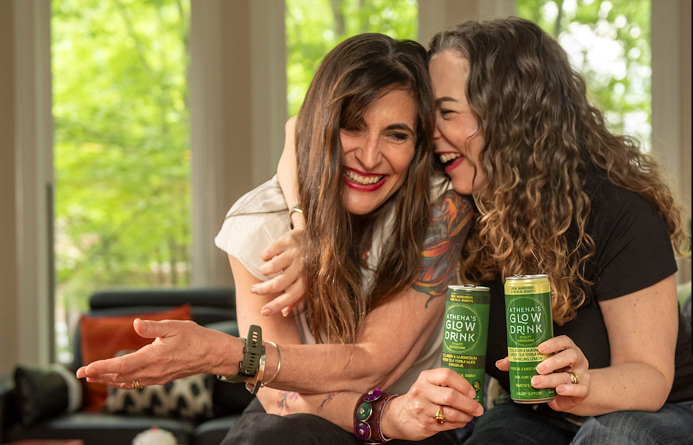 Two women sitting together holding Athena's Glow Drink cans in a casual setting.